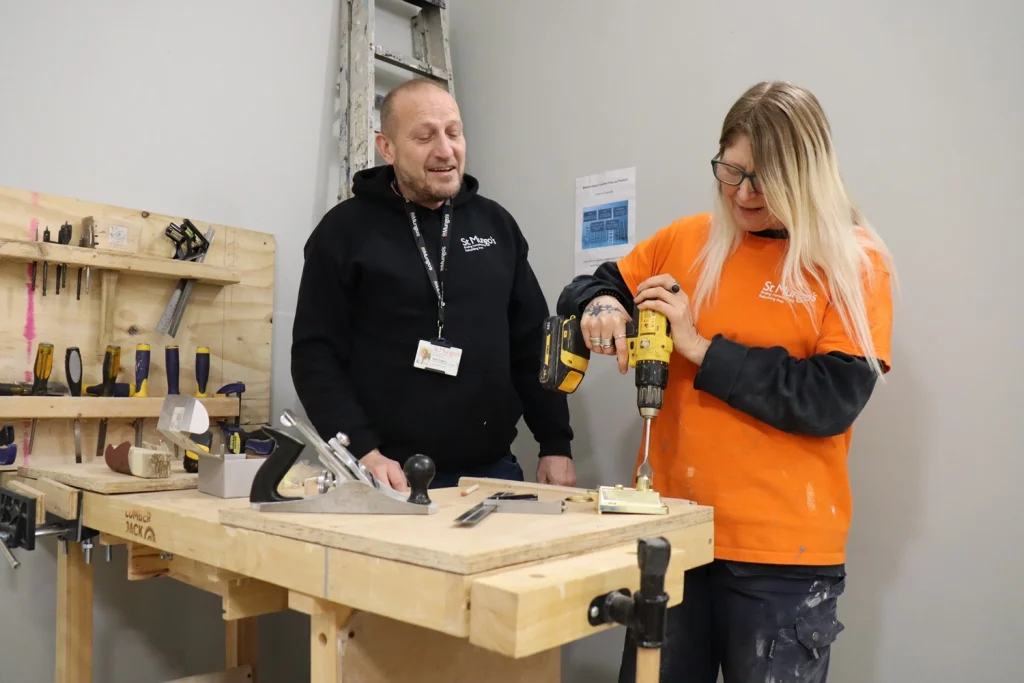 Steve Huggins, Construction Skills Manager supporting a woman (a St Mungo's client) installing hardware into a wooden frame.